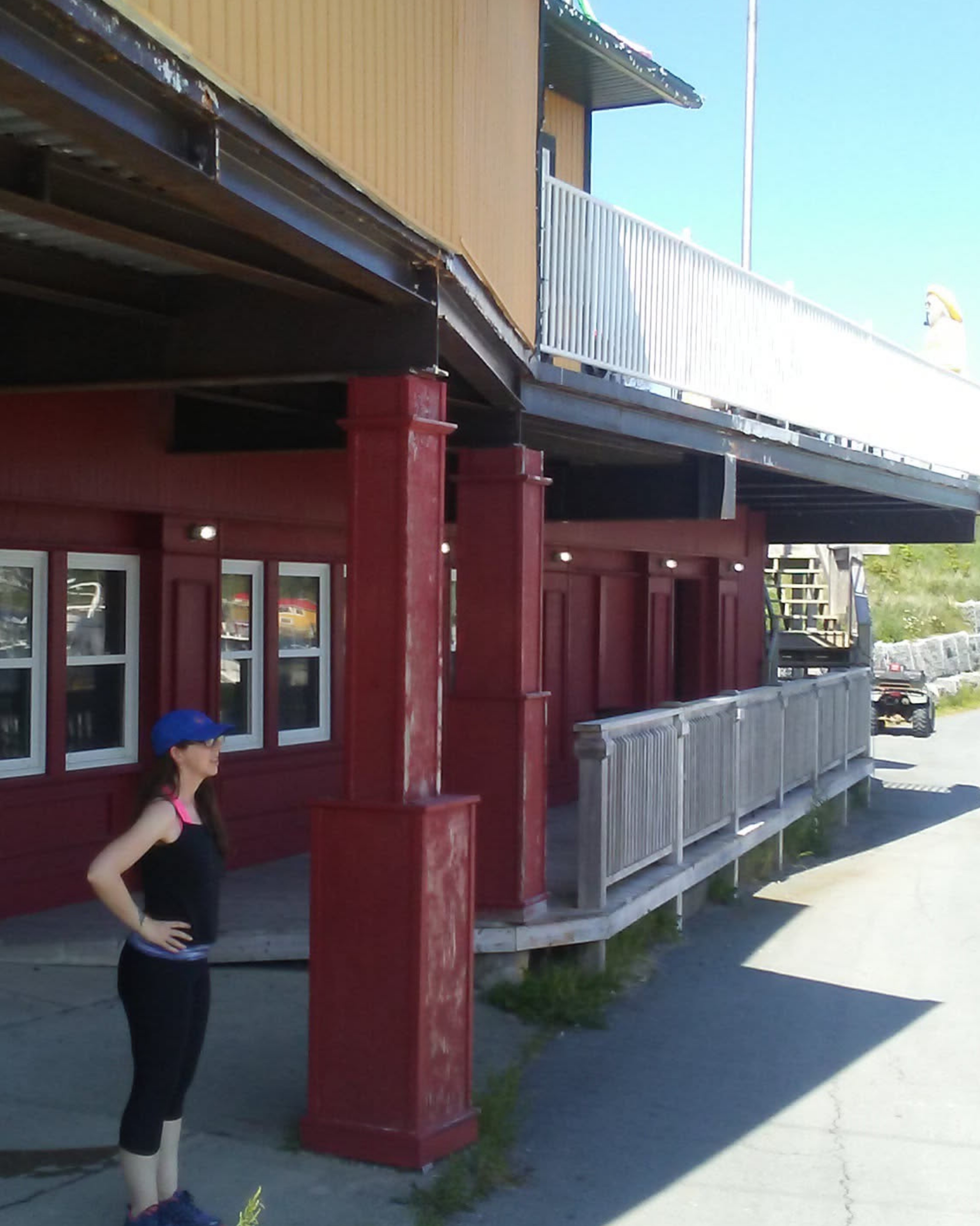 A red building wih a long veranda on a road with a stone retaining wall. A woman dressed to go for a run stands near the entrance of the building with her hands on her hips.