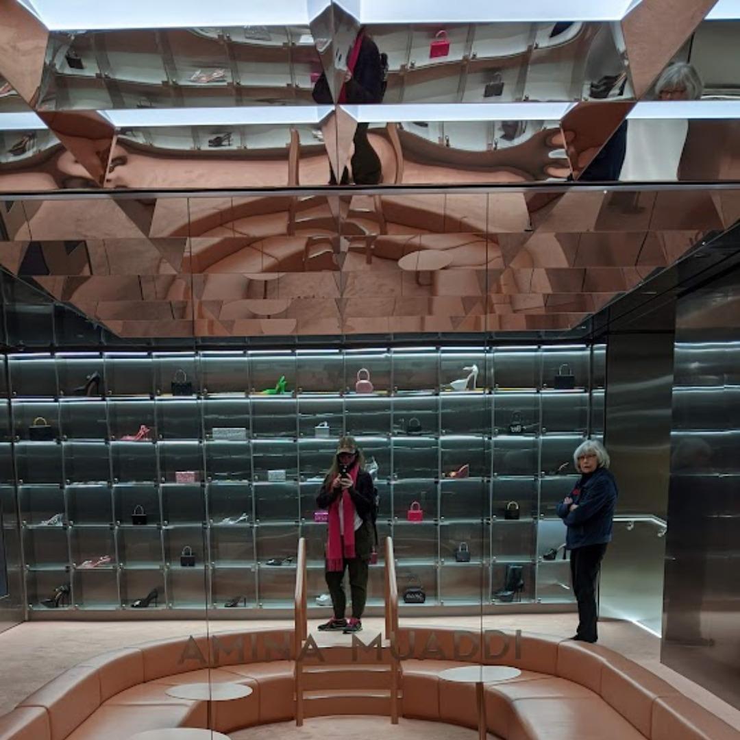 Two women in the Amina Muaddi lounge at Herrods Department store.  There is a large oval shaped seating area set in the floor, with round tables.  Behind them is a wall display of shoes and purses.  The ceiling is a mirror, so there are funky looking reflections in the upper portion of the image.