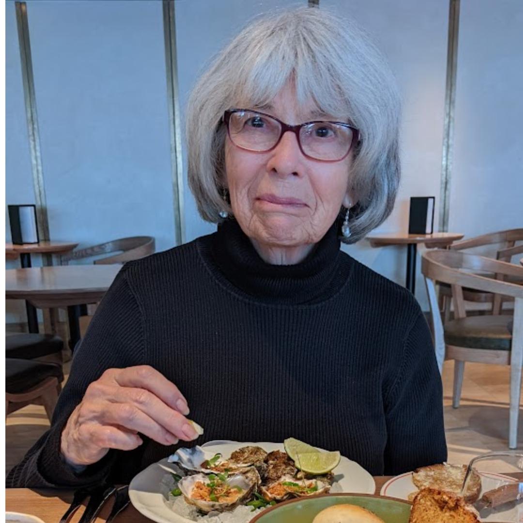 A white woman with white hair and glasses wearing a black turtle neck enjoying a plate of oysters in an art gallery restaurant.