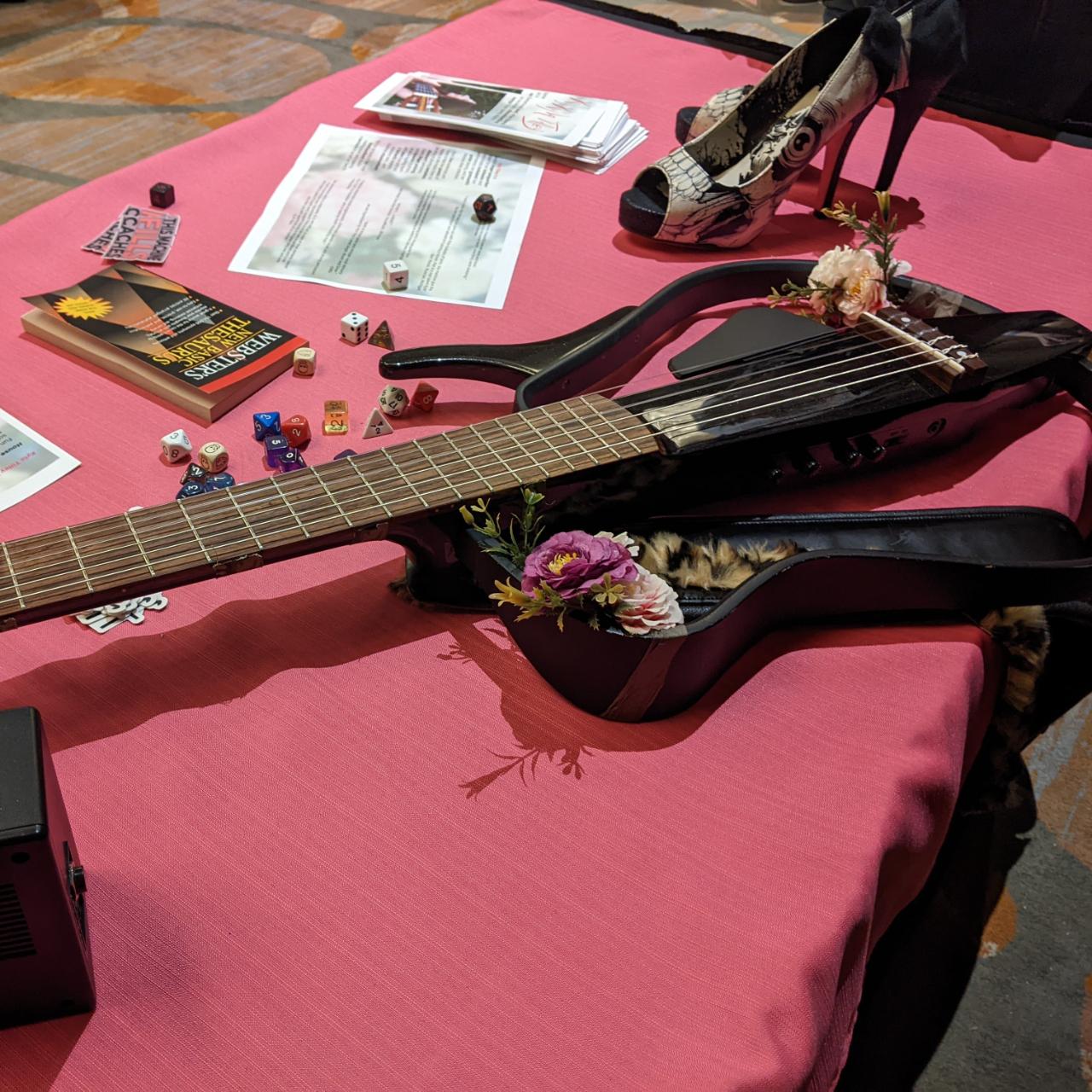 A top view shot of a singer-songwriter's booth at a trade show.  There is a lot of pink.  Pink table cloth, gothy pink soled high heels.  A handfull of D&D dice and flyers on the table, and a Black Yamaha Silent Classical Guitar with flowers attached to it. 