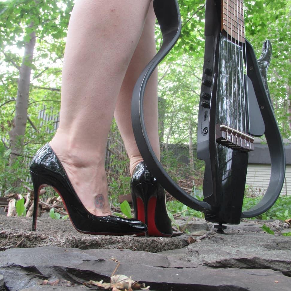 A white woman's calves in a pair of black shiny Louboutins.  She's standing on a rock patio in a verdant back yard and there is a Yamaha silent classical next to her feet.