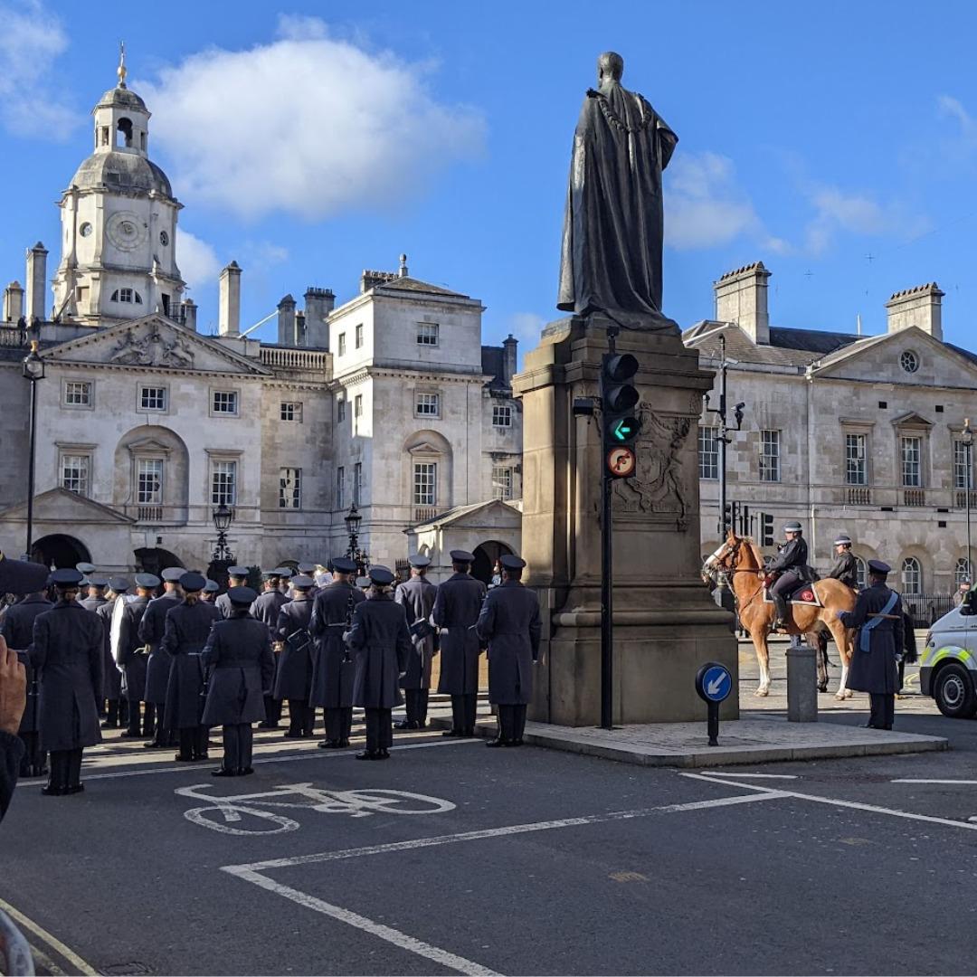 A military band from the back view standing in front of an imposing white stone building next to a statue, a bike lane, and a set of traffic lights.  Off to the side you can see more military on horse back.  They have just finished playing God Save the King.