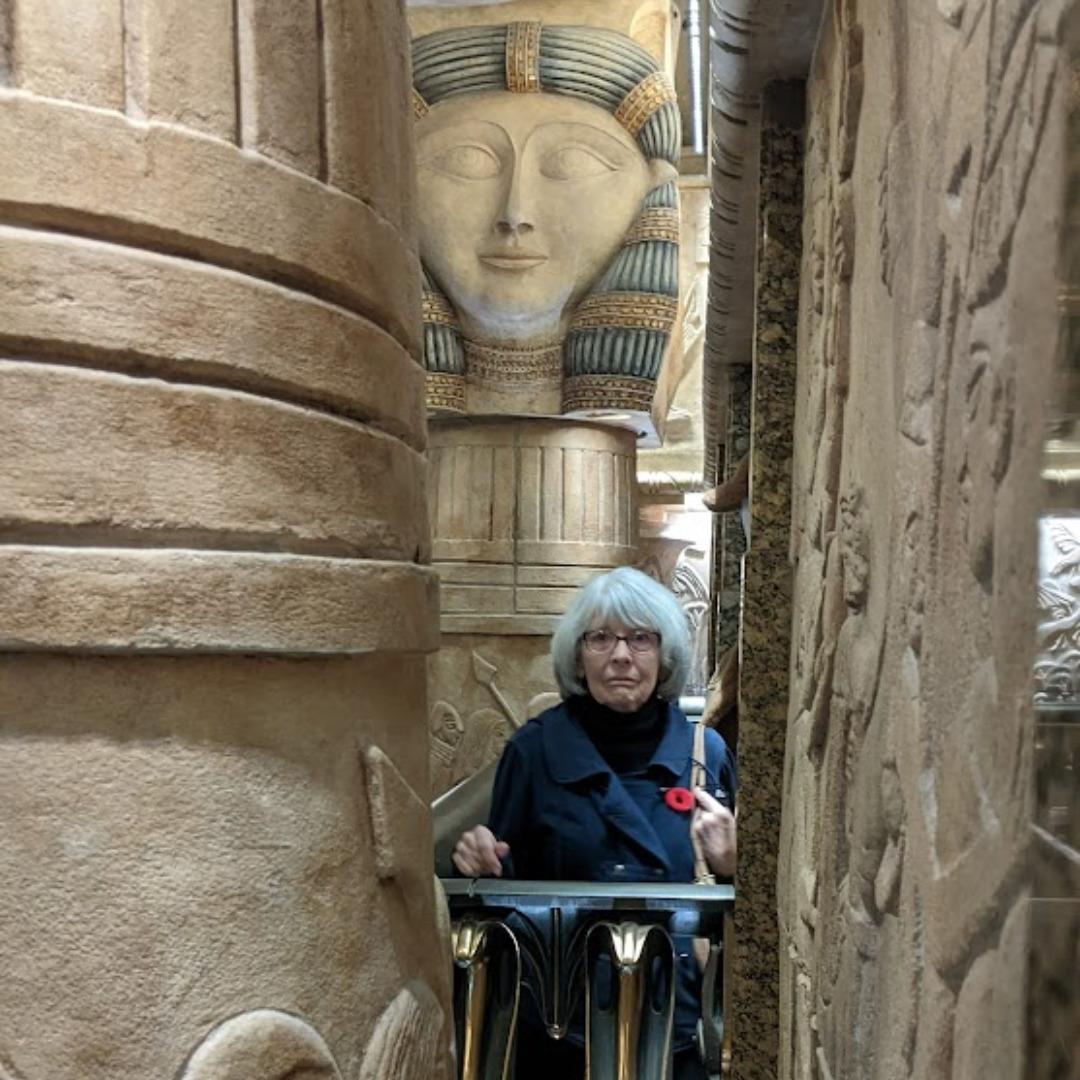 A white woman with white hair and a blue coat standing very still amongst the ancient Egyptian inspired stone sculptures decorating Herrods Department store in London.