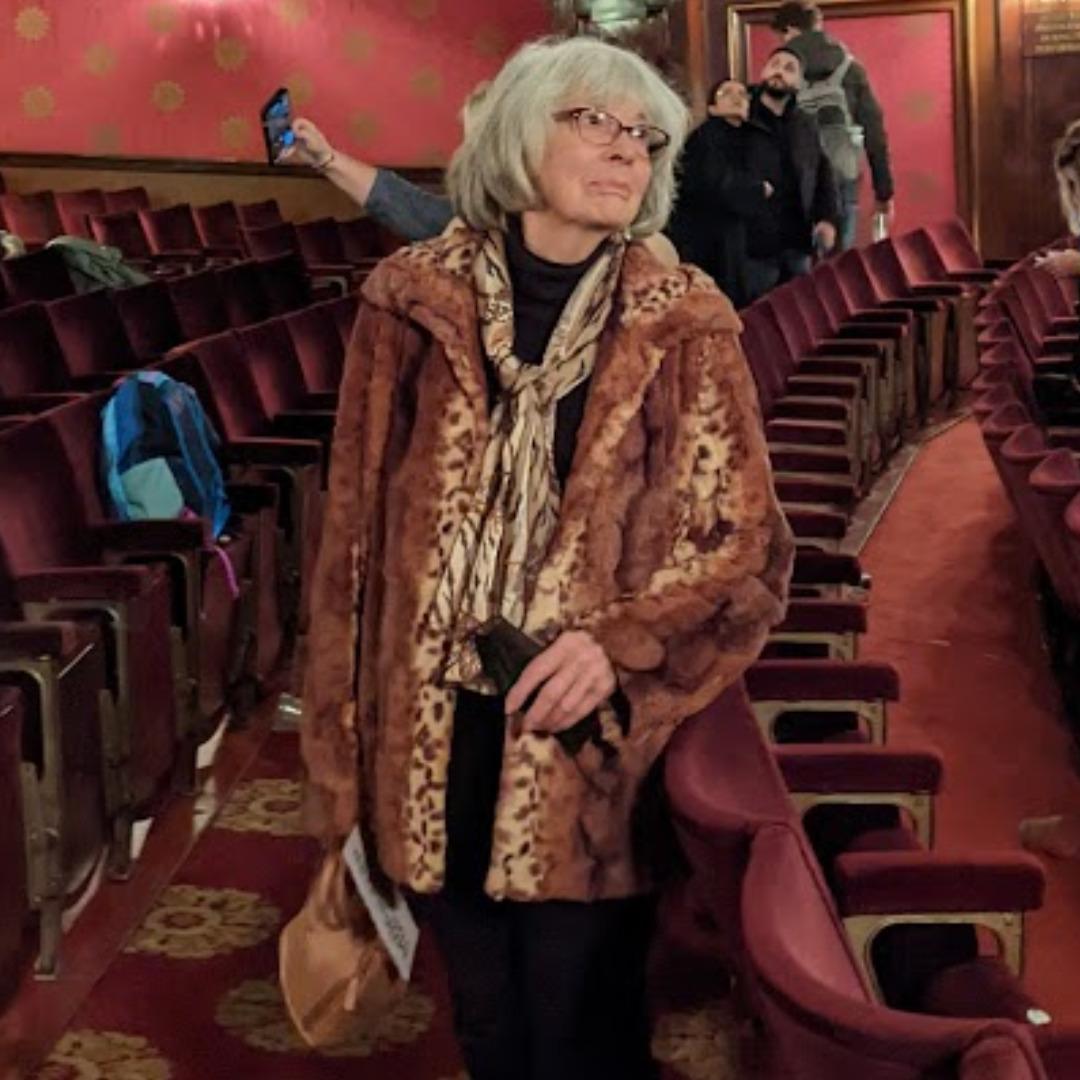 A white woman with white hair wearing a fabulous fun fur animal print coat, leaning casually in a theatre at the end of a play, looking off into the distance.