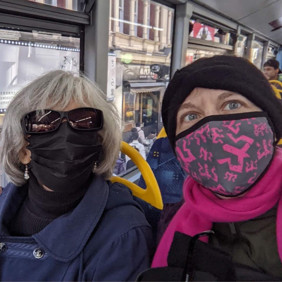 Two women riding a bus.  One has white hair, big sunglasses, and is wearing a blue coat and a black face mask.  The other is wearing a grey and pink face mask, a green coat.