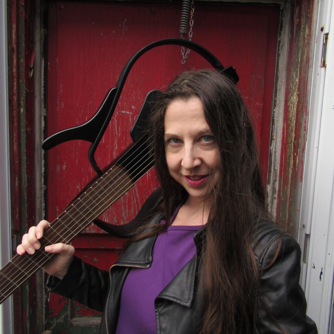 Head shot of Kyla Tilley. She's a white woman with long brown hair looking directly into the camera.  She's smiling, but also somewhat smirking.  She's wearing a purple dress ati a black leather jacket.  She's standing in front of an old red door, and holding a Yamaha Silent Classical guitar over her shoulder.