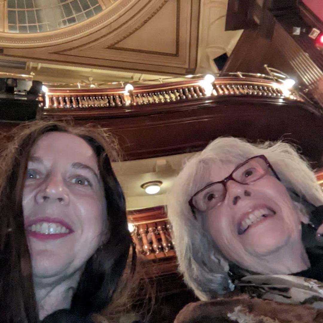 A selfie of a mother and daughter in St. Martin's Theatre, taken so that the ceiling and balconies are present.  They are both white women.  The daughter has long brown hair and is wearing pink lipstick, the mother has white hair and glasses and looks very happy to be at the theatre.