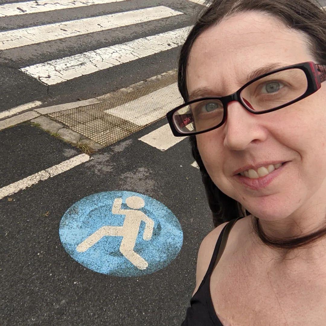 A white woman with glasses next to a sign on some ashpault that clearly denotes this as a path for runners.