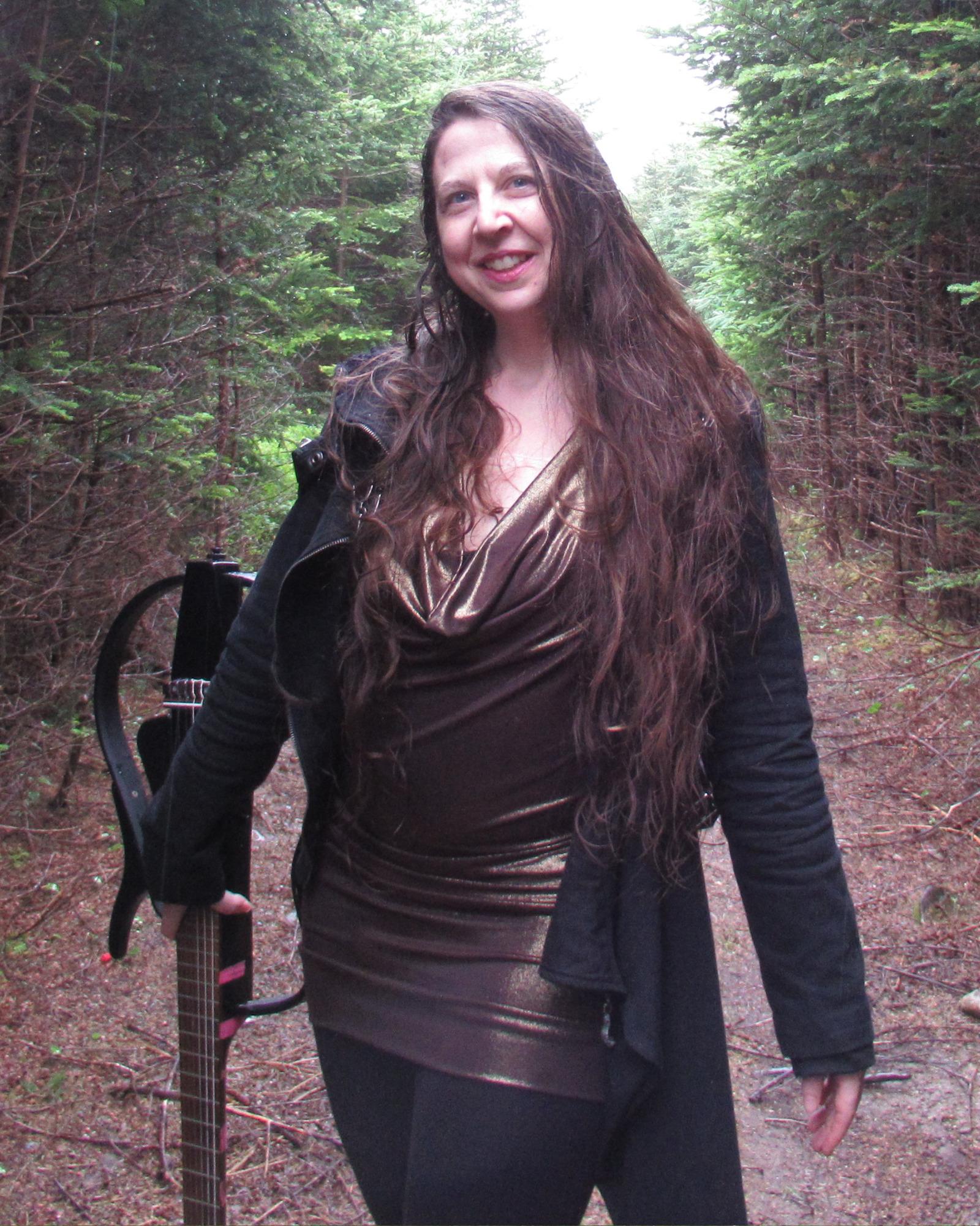 A woman with long brown hair wearing a bronze gown and a goth coat in the woods holding a futuristic looking guitar.  And smiling.